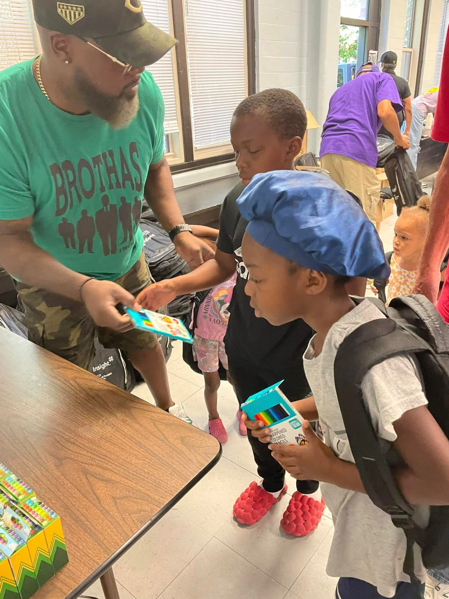 Mentor in a Brothas Inc. shirt handing colored pencils and supplies to two young boys with new backpacks