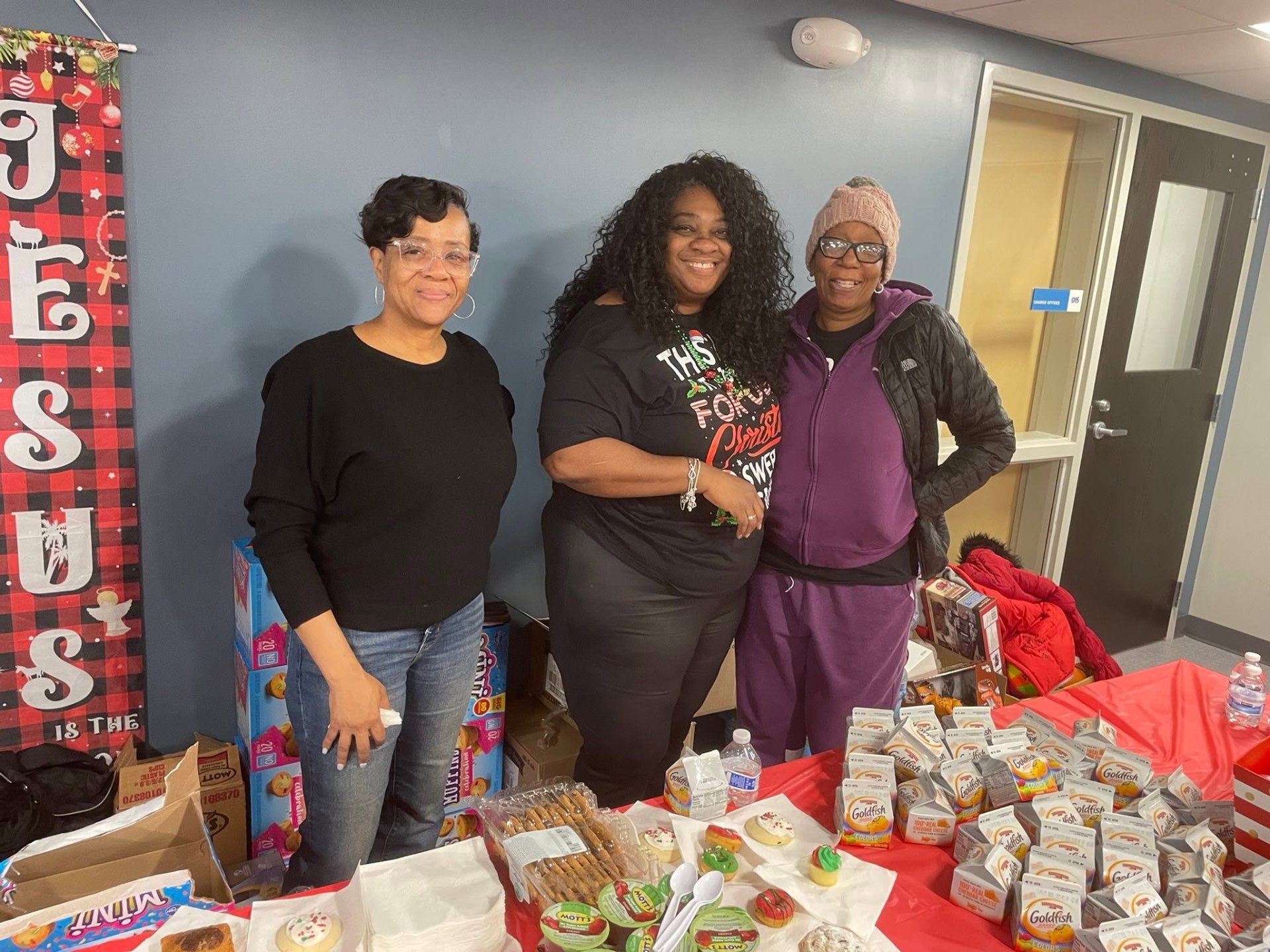 Three volunteers posing behind a table full of Goldfish, cookies and snacks at Brothas Inc. Jingle Jam 2025