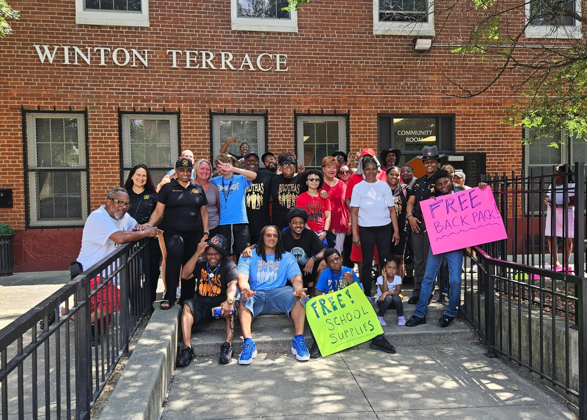 Large group of Brothas Inc. volunteers, deputies and neighbors posing with FREE BACKPACK signs outside Winton Terrace