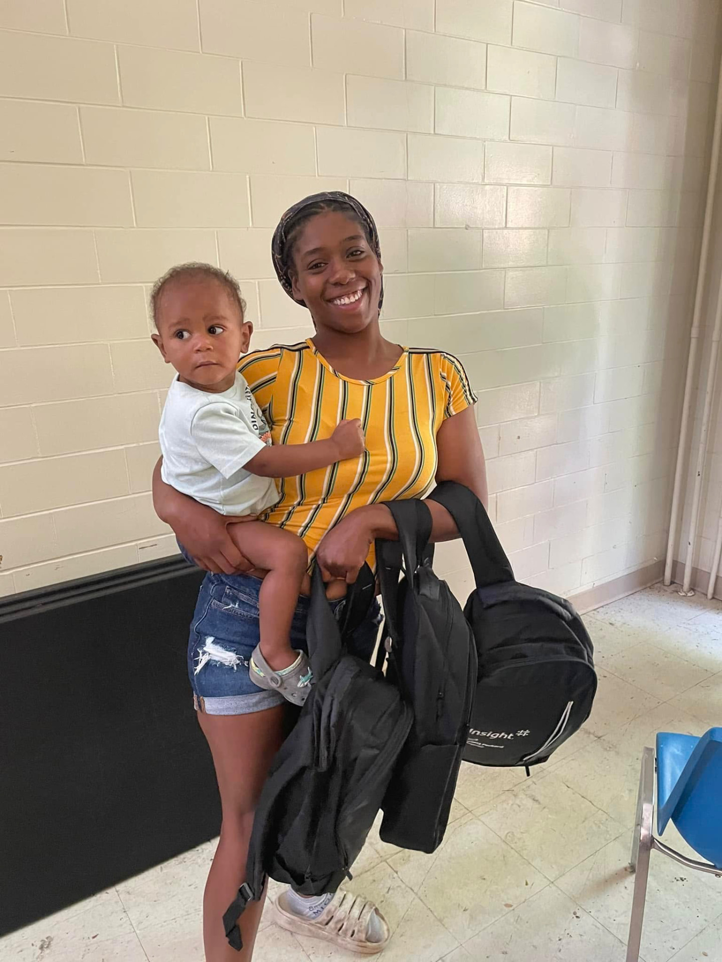 Smiling mother holding her toddler and two new backpacks at the 2024 Backpack Giveaway