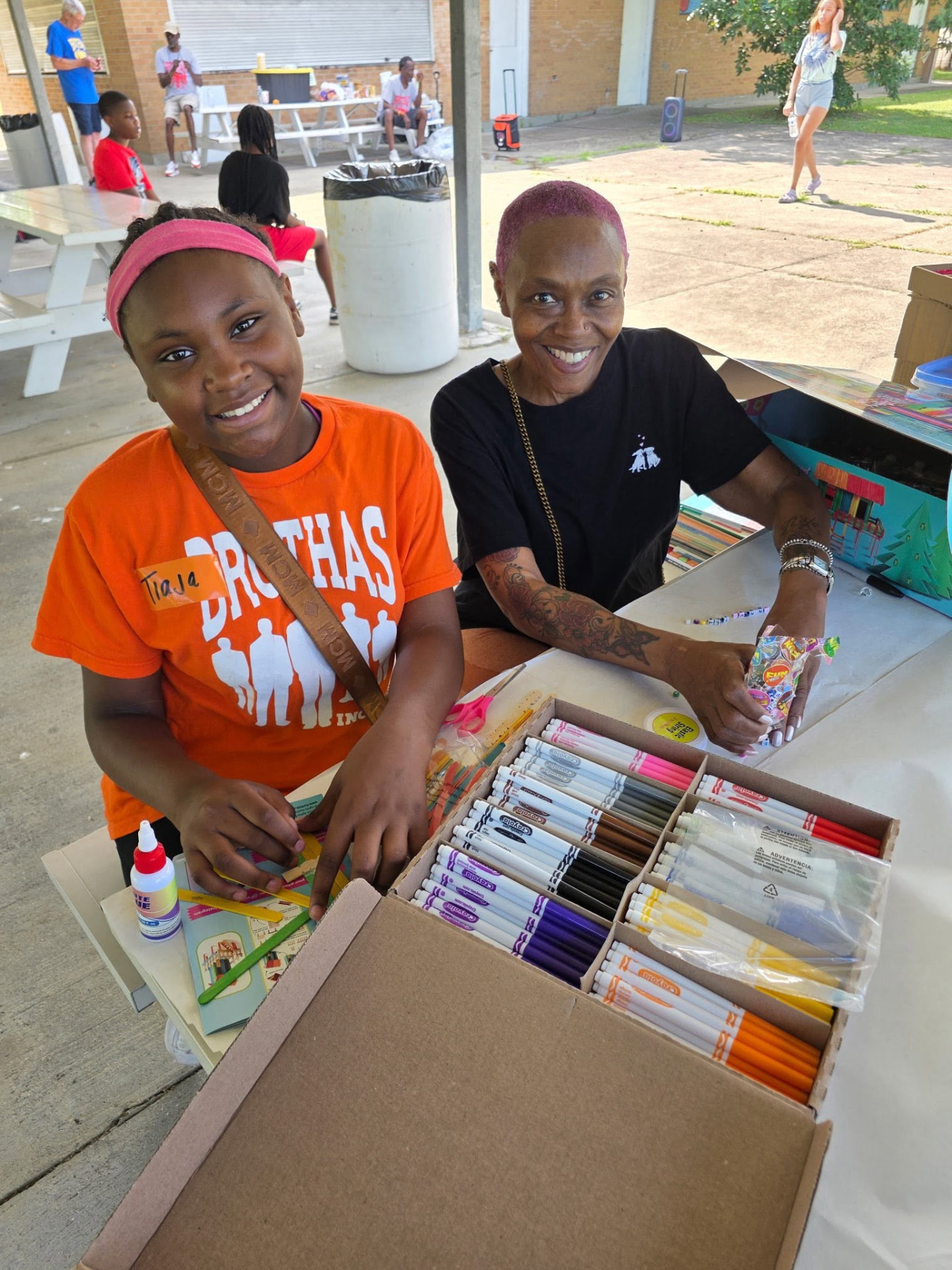 A young girl in a Brothas Inc. shirt and a volunteer smiling at the craft supplies table