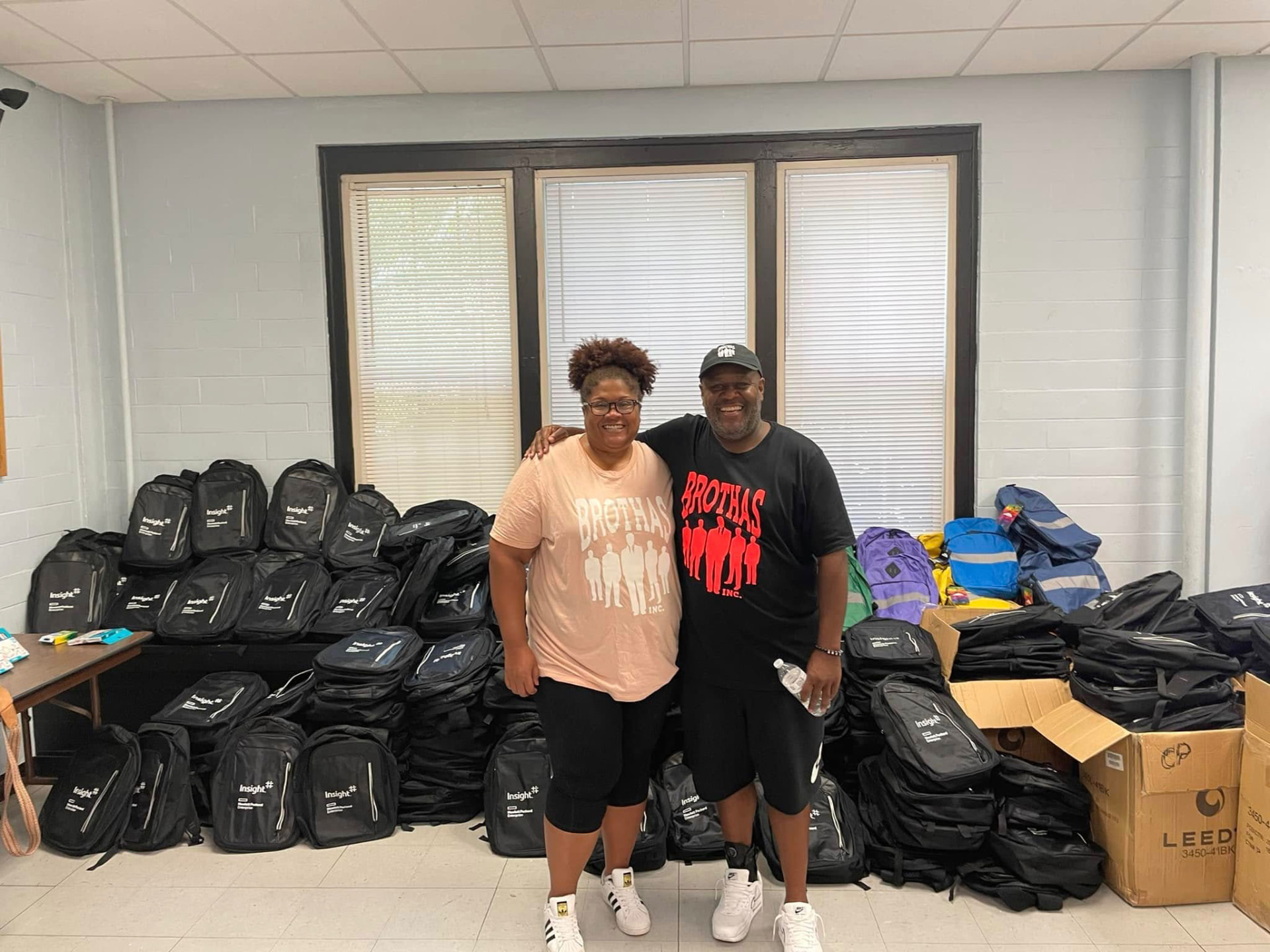 Two Brothas Inc. volunteers smiling together in front of stacks of donated backpacks at the 2024 Backpack Giveaway
