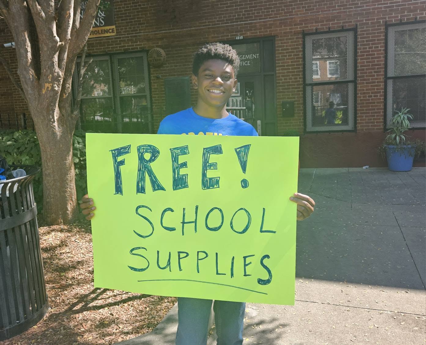 A teen volunteer holding a bright green FREE SCHOOL SUPPLIES sign at the 2025 Backpack Giveaway