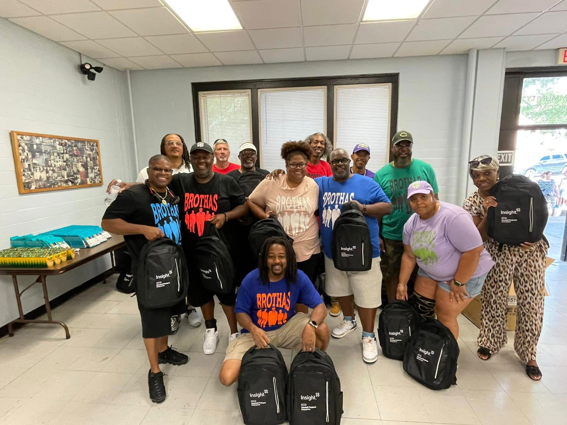 Large group of Brothas Inc. volunteers in Brothas shirts holding backpacks and smiling together at the 2024 Backpack Giveaway