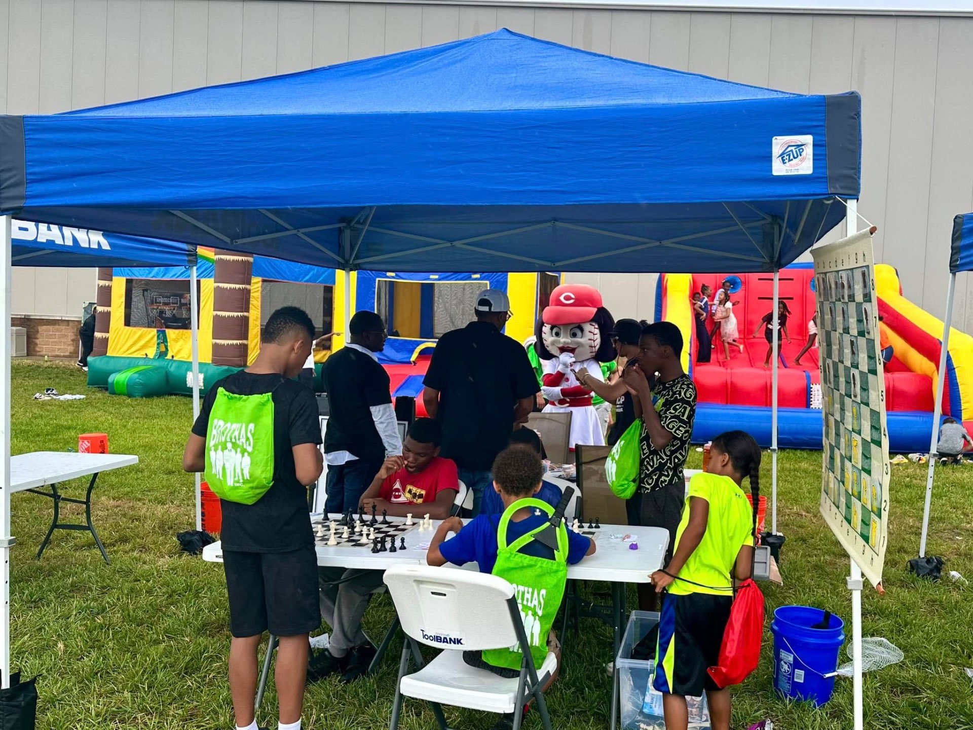 Kids playing chess under a tent with bounce houses and Rosie Red mascot at 2nd KidsFest