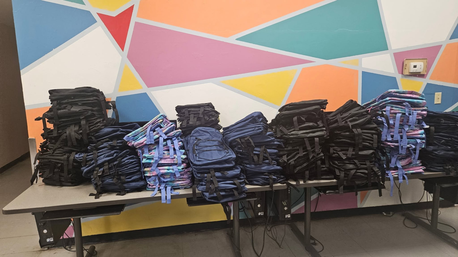 Tables stacked with dozens of backpacks in front of a colorful mural at the 2025 Backpack Giveaway