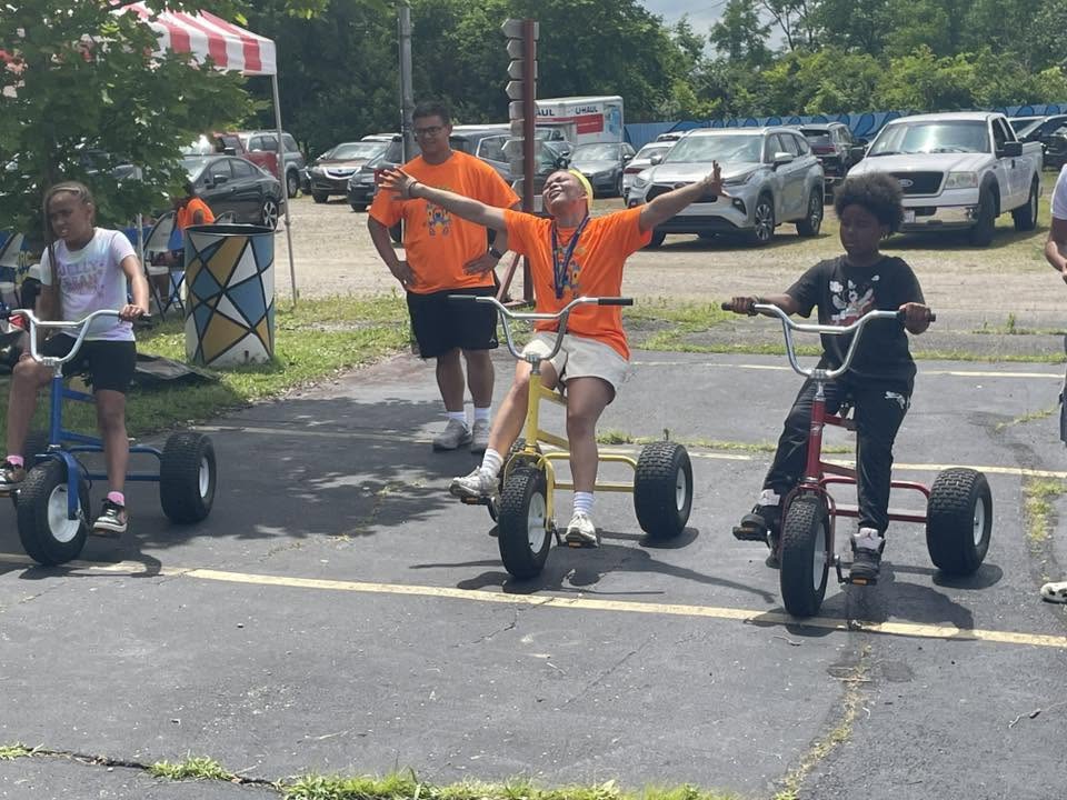 Kids riding big-wheel trikes with a mentor cheering them on at 3rd KidsFest