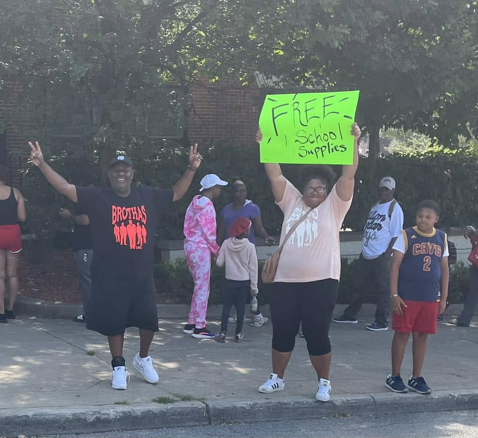 Volunteers on the sidewalk holding a bright green Free School Supplies sign at the 2024 Backpack Giveaway