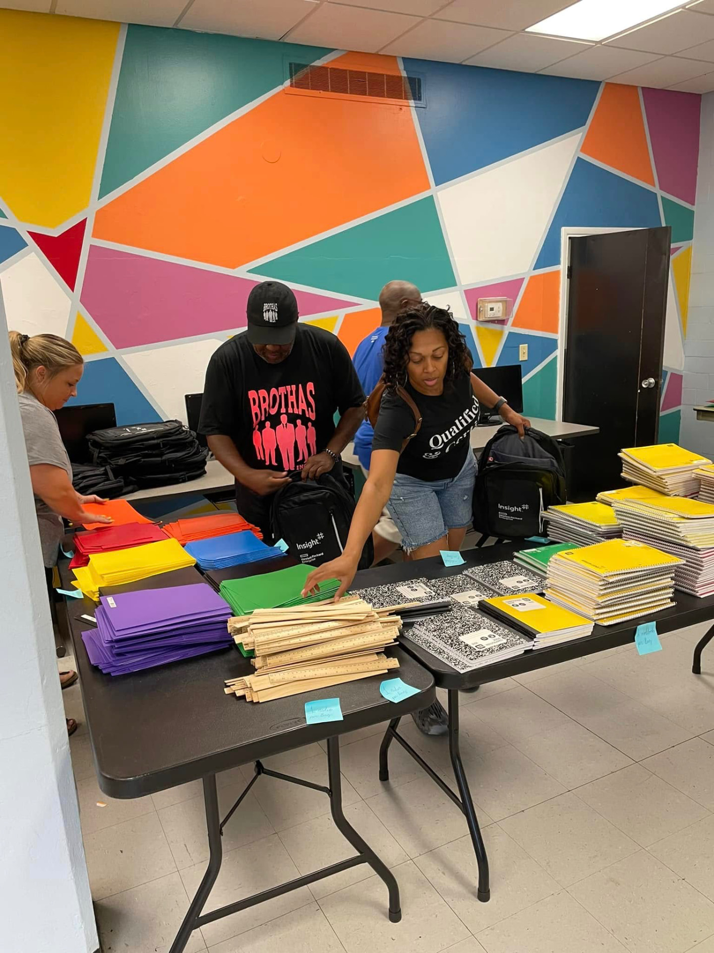 Volunteers filling backpacks with folders, notebooks and rulers in front of a colorful mural at the 2024 Backpack Giveaway