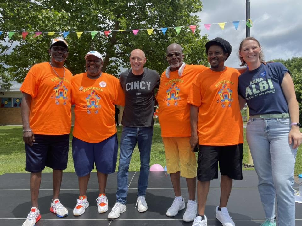 Group of mentors in orange KidsFest shirts and a CINCY tee posing with Council Member Anna Albi on stage
