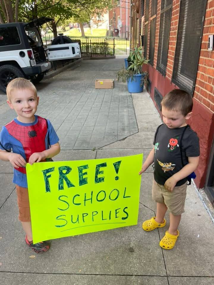 Two young boys holding a bright green FREE SCHOOL SUPPLIES sign on the sidewalk